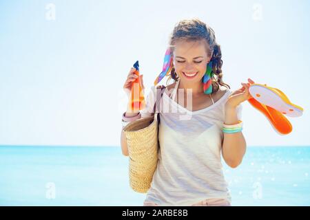 felice donna alla moda in t-shirt bianca con sacchetto di paglia sulla spiaggia, infradito arancione e bottiglia di crema solare sulla riva del mare. Foto Stock