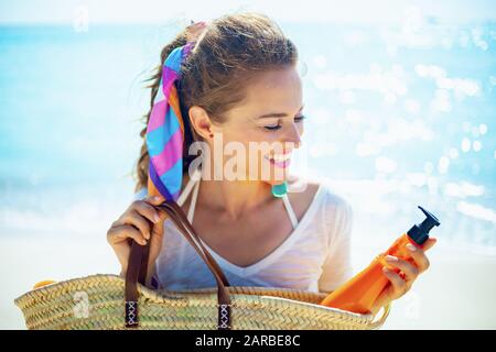 felice donna alla moda in t-shirt bianca sulla spiaggia portando bottiglia spf arancione fuori dalla borsa di paglia della spiaggia. Foto Stock