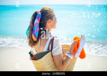 Visto da dietro donna moderna in t-shirt bianca con borsa di paglia da spiaggia, infradito arancione e bottiglia di spf che guarda in lontananza sulla spiaggia. Foto Stock