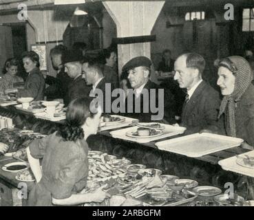Scena all'interno di un ristorante britannico, uno dei numerosi ristoranti comuni durante la seconda Guerra Mondiale che offre un pasto cotto, con dessert e una tazza di tè, al costo di circa 1 s.2d. Mentre operavano su base mensa, i pasti venivano consumati a un tavolo e permettevano a persone, compresi gli operai di guerra e coloro che avevano bombardato fuori casa o che potevano avere difficoltà a cucinare o a preparare un pranzo al sacco a casa, un pasto nutriente e conveniente, che potrebbe essere mangiato in aggiunta alle razioni in tempo di guerra. La fotografia mostra i clienti che fanno la coda al bancone. Foto Stock