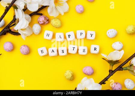 Buona Pasqua. Cubi con testo su sfondo giallo. Brunch primaverili con fiori bianchi e caramelle, uova di pasqua al cioccolato. Piatto minimalista. Foto Stock