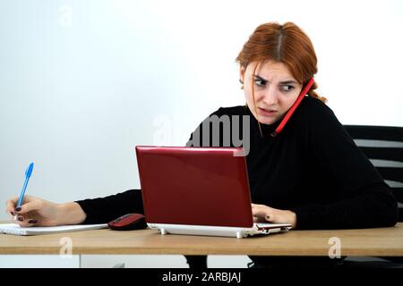 Giovane lavoratore di ufficio donna a parlare su un telefono cellulare seduto alla scrivania con computer laptop e notebook. Foto Stock