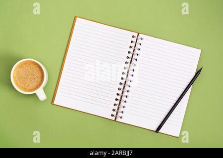 Aprire il notebook con una pagina vuota e la tazza di caffè. Piano superiore dello spazio di lavoro su sfondo verde. Piatti creativi laici. Foto Stock