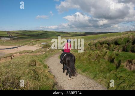 Young Female Riding a Horse on the South West Coast Path Down to the Beach of Gunwalloe Church Cove in Rural Cornwall, England, UK Foto Stock