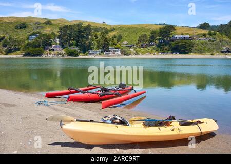 Spiaggia di ciottoli e la città di Jenner sulle colline attraverso la foce del fiume russo in California. Canoe gialle e rosse ormeggiate sulla riva. Foto Stock