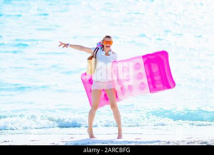 donna sorridente e sana di 40 anni in maglietta bianca e pantaloncini rosa con sacchetto di paglia sulla spiaggia che gioisce mentre tiene il materasso gonfiabile. Foto Stock