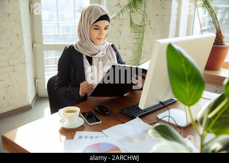 Revisione dei documenti. Ritratto di una bella donna d'affari araba che indossa hijab mentre lavora in openspace o ufficio. Concetto di occupazione, libertà nell'area di business, successo, soluzione moderna. Foto Stock