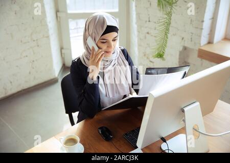 Parlare al telefono durante la revisione dei documenti. Bella donna d'affari arabo che indossa hijab mentre lavora in openspace o ufficio. Concetto di occupazione, libertà nell'area di business, successo, soluzione moderna. Foto Stock