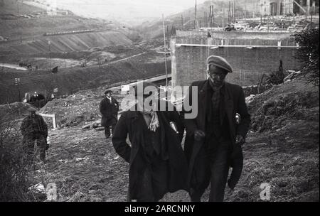 1940s, storico, lavoratori delle miniere indossando soprabiti e cappotti e con facce annerite coperte di fuliggine, camminando sulla collina dalla miniera di carbone nella valle sottostante, Merthyr, Galles, Regno Unito. Foto Stock