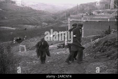 1940s, storico, lavoratori delle miniere indossando soprabiti e cappotti e con facce annerite coperte di fuliggine, camminando sulla collina dalla miniera di carbone nella valle sottostante, Merthyr, Galles, Regno Unito. Foto Stock