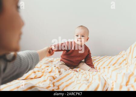 Bella bambina sul letto in un'accogliente maglione marrone sorride. Concetto di maternità e di infanzia. Adorabile bambina di sei mesi sdraiata sul male e guardando nella fotocamera. Madre tenere poca mano. Foto Stock