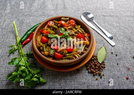 Fegato di pollo con verdure su un piatto di argilla. Pezzi di fegato, cipolla, fagiolini, pomodoro. Tela grigia testurizzata. Intorno alla piastra è verde e rosso c Foto Stock