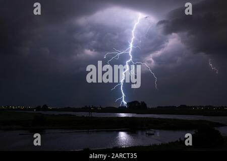 Fulmine luminoso con molte diramazioni laterali colpisce la terra in un paesaggio fluviale Foto Stock