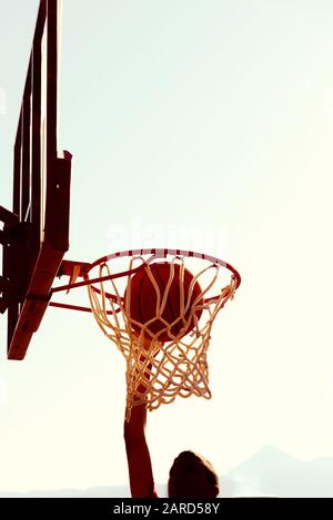 Il giovane giocatore di basket dalla silhouette sinua che salta in alto sul cerchio di basket raggiunge un punteggio con un dunk di volo al tramonto. Successo e obiettivo Foto Stock