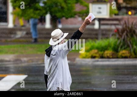 Vista posteriore dell'uomo che indossa cappello e maglietta bianca mentre mostra carta bianca per fare l'annuncio per l'evento. Su strada durante la stagione delle piogge Foto Stock