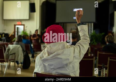 Vista posteriore della persona in cappello rosa e felpa con cappuccio seduto sulla sedia mentre solleva la mano e fa domanda al diffusore. Durante il discorso in auditorium Foto Stock