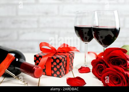 Concetto di San Valentino. Due bicchieri di vino, rose rosse, regalo, petali di rosa, bottiglia di vino e cavatappi su un tavolo di legno bianco. Messa a fuoco selettiva. Foto Stock