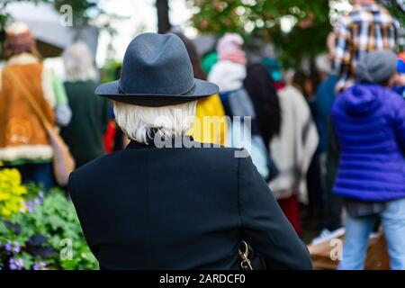 Vista posteriore della donna con cappello grigio e blazer blu mentre si guarda defocus folla in piedi in evento durante il festival Foto Stock