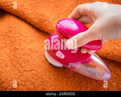 Cura dei vestiti. Rasoio per pelucchi o rasoio in tessuto o smacchiatore di pelucchi nella mano femmina. Donna che rimuove il lanugine sul rivestimento di lana arancione con defuzzer elettrico portatile. Foto Stock
