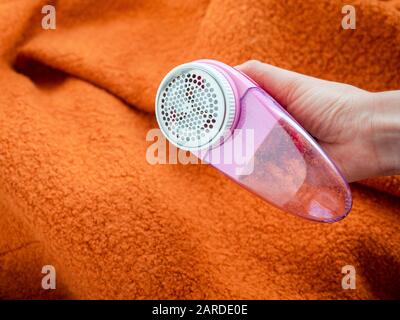 Cura dei vestiti. Rasoio per pelucchi o rasoio in tessuto o smacchiatore di pelucchi nella mano femmina. Donna che rimuove il lanugine sul rivestimento di lana arancione con defuzzer elettrico portatile. Foto Stock