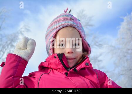 Ritratto di giovane ragazza in abiti invernali e colorato cappello di lana, pronto a lanciare palla di neve, contro cielo blu e bianco, congelati alberi bokeh Foto Stock