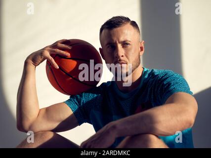 Ritratto del giocatore di pallacanestro che tiene la palla sulla spalla e seduto vicino alla parete con le ombre dalla finestra Foto Stock