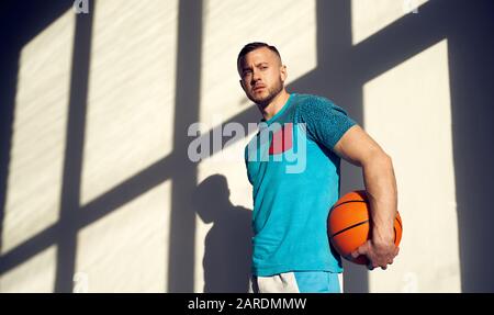 Giovane atletico, giocatore di pallacanestro che tiene la palla e sta accanto alla parete con le ombre dalla finestra Foto Stock