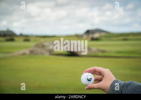 Saint Andrews, Scozia, Regno Unito - 20 giugno 2019: Sfera di golf di St. Andrews tenuta in una mano caucasica con una vista distante del ponte di Swilcan al foro 18th di Foto Stock