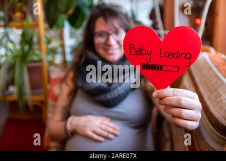 Una vista selettiva di fuoco di una donna incinta felice che tiene un messaggio rosso a forma di cuore che dice il caricamento del bambino con la barra di avanzamento. Copiare lo spazio a sinistra Foto Stock