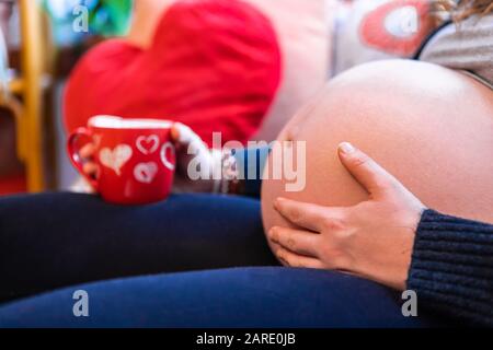 Una vista selettiva di fuoco di una donna gravida pesante con grande tummy gonfio rilassante all'interno del giorno delle valentines, con tazza blurry di tè in background Foto Stock