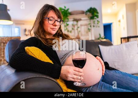 Ritratto di una donna gravida seduta su un divano in casa con un bicchiere di vino rosso, ignaro dei rischi della sindrome dell'alcol fetale. Copiare lo spazio a destra Foto Stock
