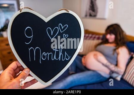 I genitori in attesa condividono il messaggio creativo su un segno a forma di cuore, la donna gravida pesante è vista sfocata nel letto a nove mesi di fase, preparandosi per la nascita Foto Stock