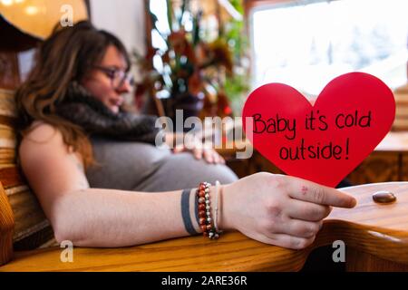 Una vista selettiva di fuoco di una donna gravida che tiene un messaggio rosso di cuore che dice al bambino il relativo freddo fuori, preparando per il parto a San Valentino Foto Stock