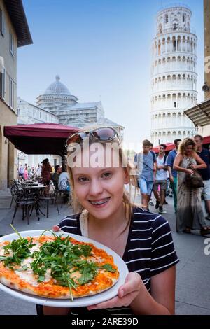 Ragazza adolescente con capelli biondi, occhi di nocciola e bretelle dentali tiene una pizza in un caffè all'aperto con la Torre Pendente di Pisa sullo sfondo. Foto Stock