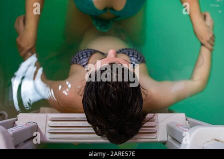 Vista ad alto angolo della donna incinta appoggiata sulla scala in bikini mentre la donna terapista tiene la mano e dà idroterapia in piscina Foto Stock