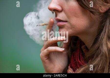 Closeup taglio corto di metà inferiore del viso della donna caucasica fumo mano rotolato marijuana comune. Il fumo di cannabis penetra dalla bocca di una donna edgy Foto Stock