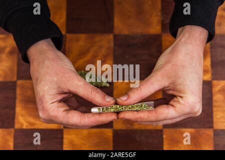 Colpo di Closeup delle mani che pizzicava una carta da rotolamento riempita di cannabis macinata che si prepara a rotolarla in una giuntura. Germoglio di erbaccia sulla tabella a scacchi qui sotto. Foto Stock