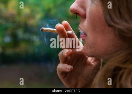 Closeup shot di metà inferiore della donna caucasica viso fumo mano rotolò marijuana comune. La sigaretta di cannabis appena illuminata tenuta tra le sue due dita. Foto Stock