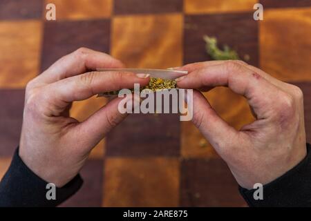 Colpo di closeup superiore delle dita che comprimono una carta arrotolabile riempita di cannabis a terra che comincia a torcerla in un giunto sopra una tabella a scacchi. Foto Stock