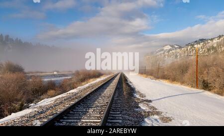 Un tratto luminoso e soleggiato di binari ferroviari che corrono lungo il fiume Clark Fork svanisce nella fredda nebbia invernale. Bearmouth, Montana Foto Stock