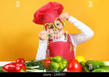 Un bambino allegro nel cappello di uno chef ha divertimento con le verdure e tiene il pepe davanti ai suoi occhi, come se in bicchieri. Piccola ragazza siede ad un tavolo su un bac Foto Stock