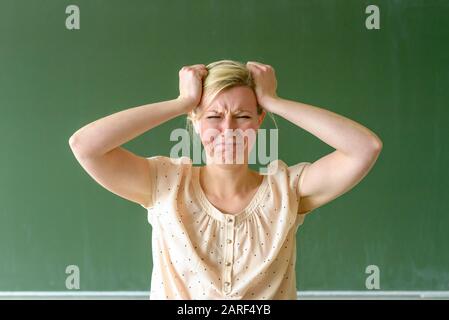 Arrabbiato frustrato insegnante di scuola strappando ai capelli con le mani e avvitando il suo volto in piedi di fronte a una lavagna in classe Foto Stock