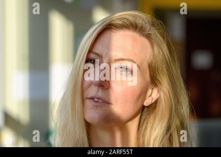 Donna bionda scettica che guarda sospettosamente alla fotocamera con un'espressione meditata vicino a una finestra con luce solare riflessa sul suo viso Foto Stock