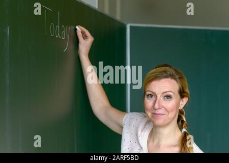 Primo piano ritratto di insegnante femminile in classe scrivere sulla lavagna Oggi lezione e guardare indietro i suoi studenti con il volto amichevole Foto Stock