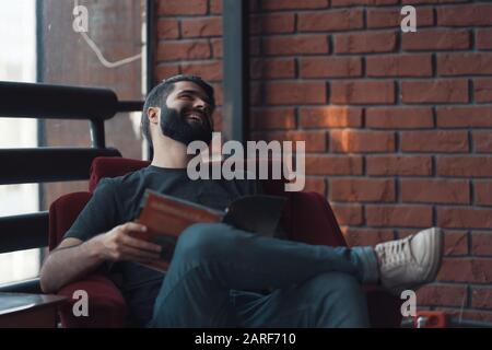 Ritratto bello uomo barbuto indossando un abbigliamento casual. Uomo seduto in poltrona rosso moderno studio loft, leggendo fumetti e rilassante Foto Stock