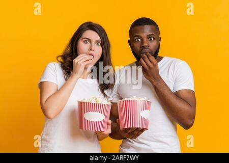 Ritratto di coppia interracial dazed che mangia popcorn con espressione di faccia spaventata Foto Stock