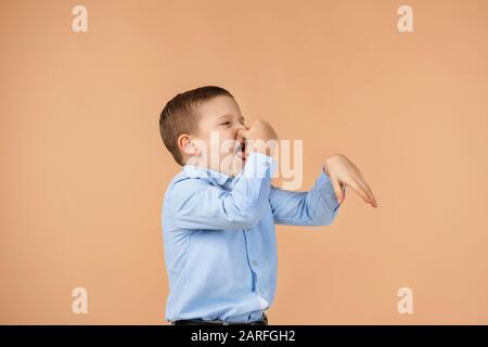 bambino piccolo ragazzo in camicia blu che fa gesto odori cattivo su sfondo beige. espressione facciale. bambino copre il naso con la mano, odori qualcosa di terribile Foto Stock