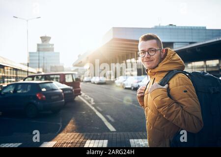 Sorridente giovane uomo con zaino sul parcheggio aganist terminal dell'aeroporto edificio al tramonto. Foto Stock