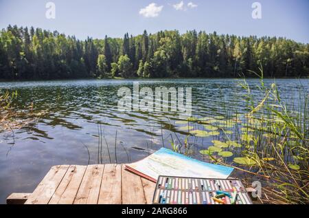 Set di pastelli pastello con disegno si adagia su un molo di legno presso un lago forestale in una luminosa giornata estiva - plein air scetching concetto Foto Stock