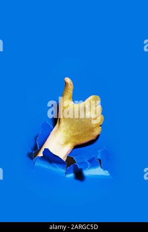 Mano con glitter dorato rompere attraverso la carta con il pollice su sfondo blu. Foto Stock